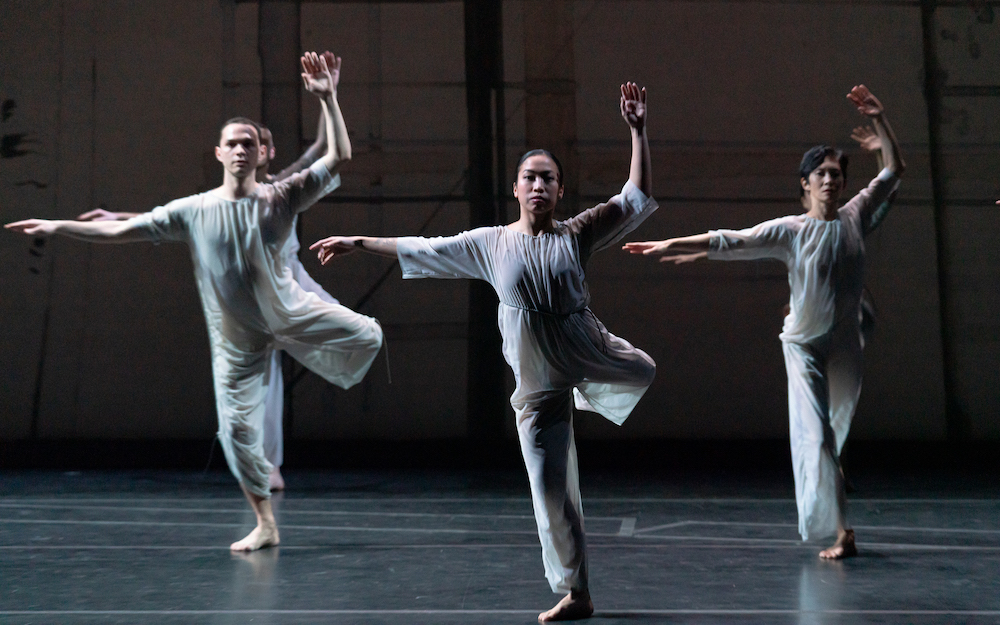 five dancers in the flowing, grey-blue jump suits face us directly, squarely, with purpose. They balance on one leg with the other bent behind them and lifted 90 degrees to their back.